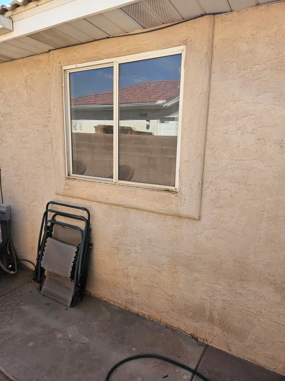An old white aluminium sliding window in a tan stucco wall on a covered patio, with a folded chair leaning beside it