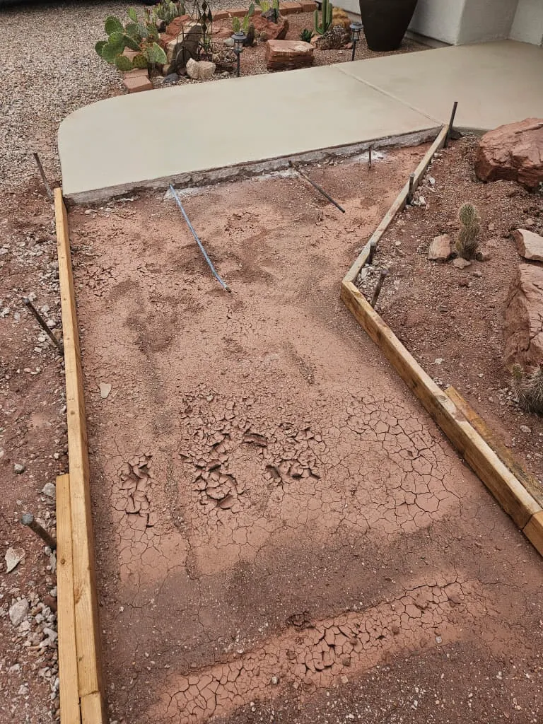 A formed and staked walkway subgrade of cracked red clay butting an existing concrete slab, with a rebar strand and a cactus bed beyond