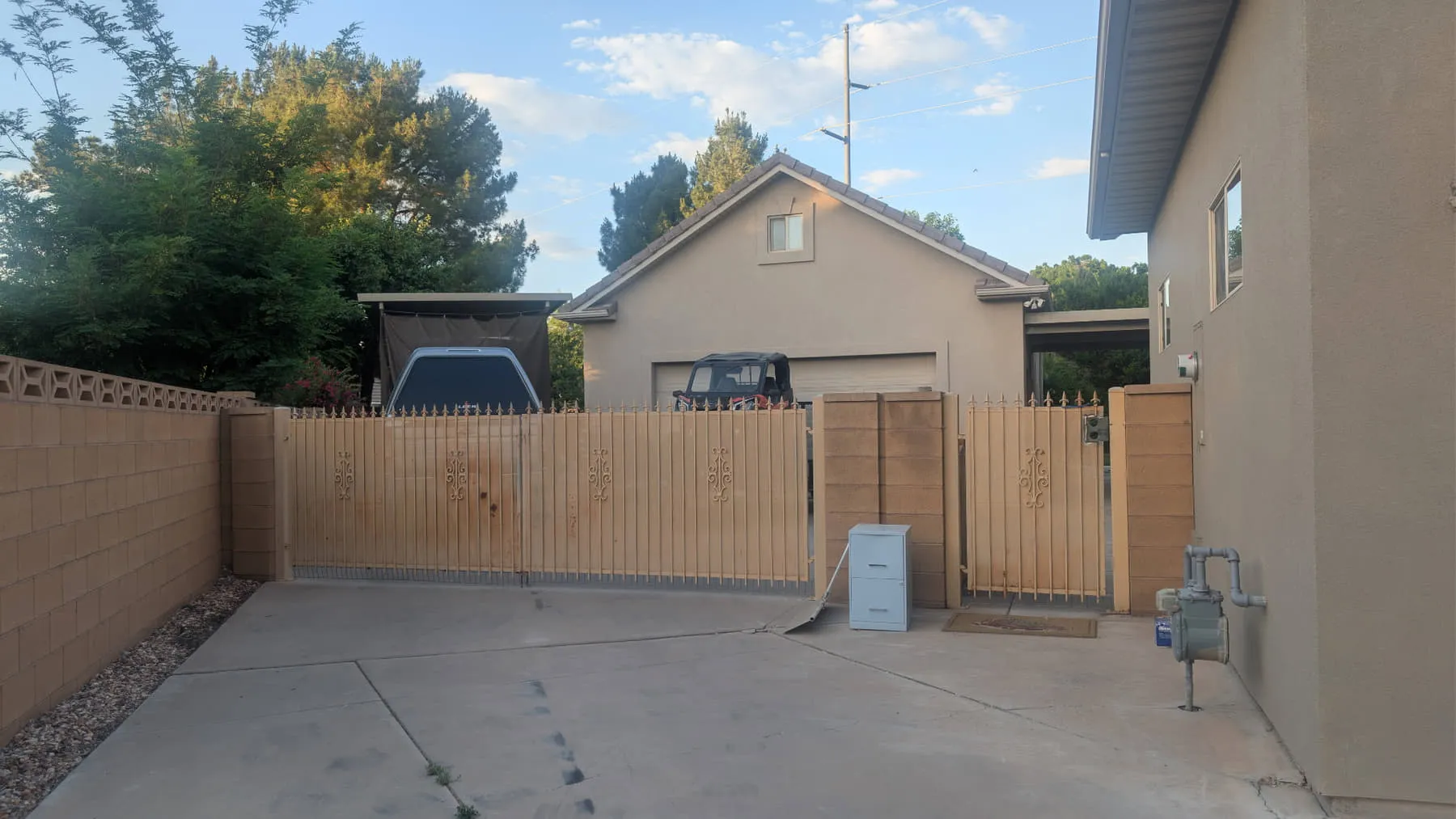 A faded tan wrought-iron double driveway gate with spear tops closed across a concrete driveway between tan block pilasters