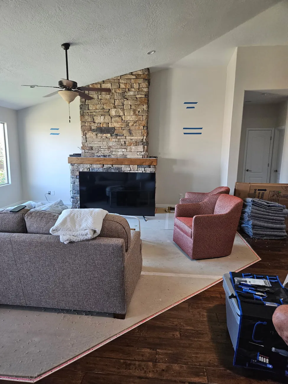 A great room with blue painter's tape marking shelf positions on the walls flanking a stacked stone fireplace, furniture pushed aside