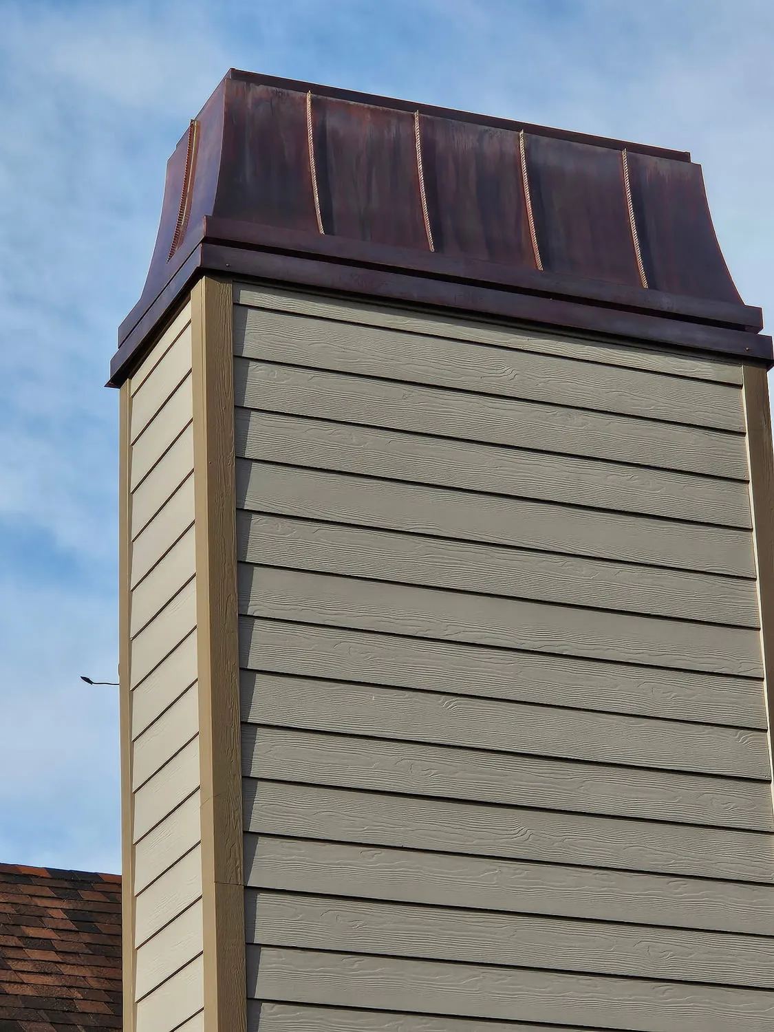 A chimney chase clad in fibre-cement lap siding with tan corner boards, topped with a custom copper shroud against a blue sky