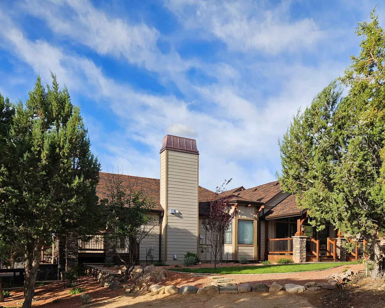 A wide view of a ranch style home showing the finished siding chimney chase and copper cap above stone porch piers and cedar rails