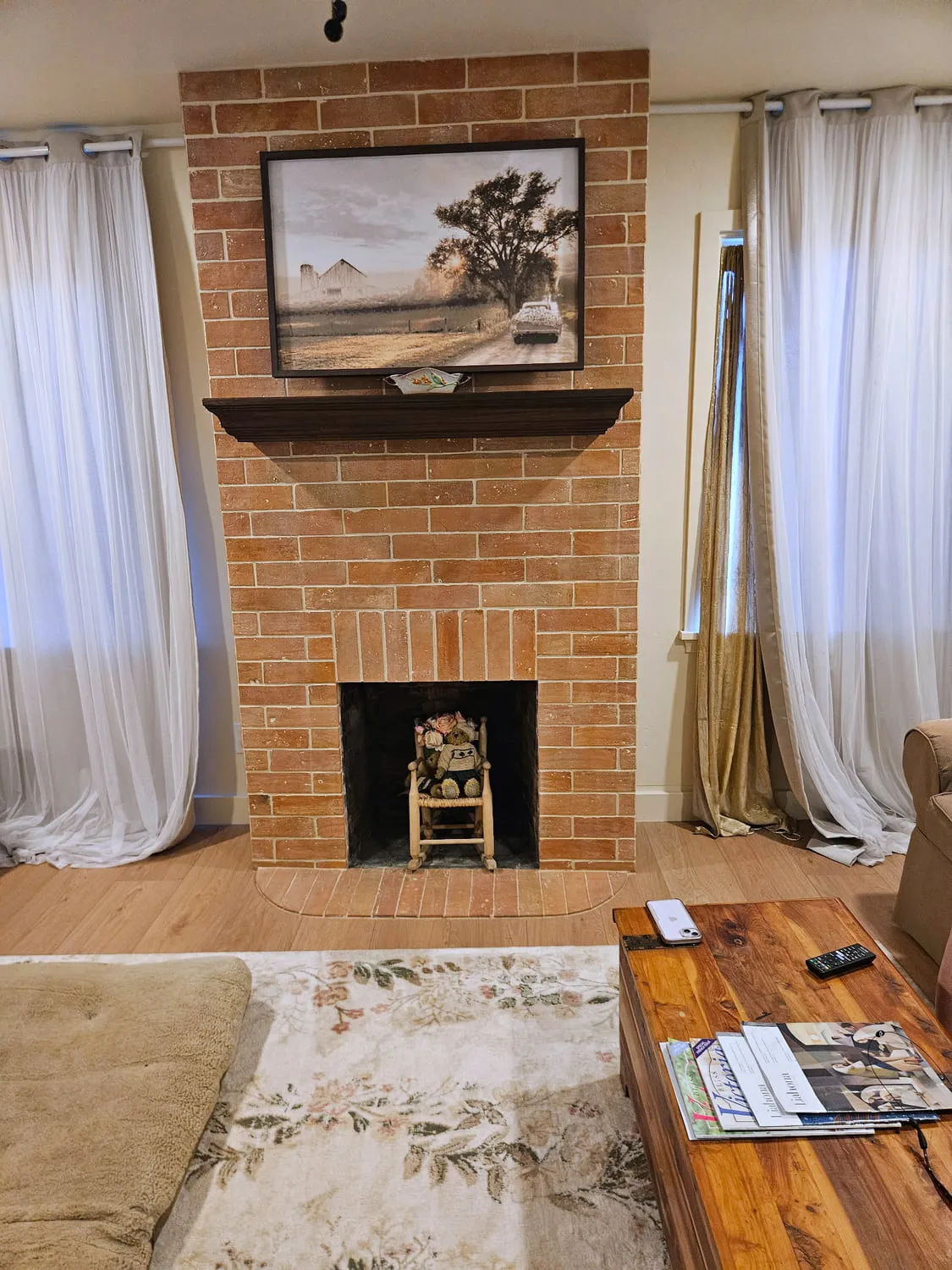 A finished full-height thin brick fireplace surround and hearth with a stained mantel, framed barn art above and sheer curtains either side