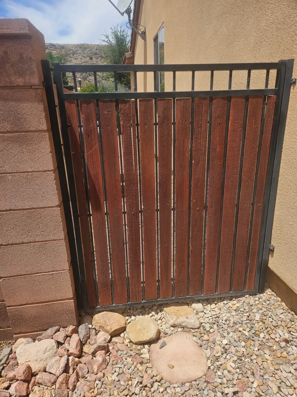 A black steel frame side gate infilled with cedar pickets freshly stained dark red, hung between a block pilaster and a stucco wall