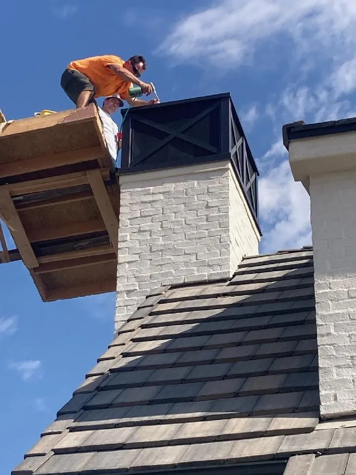 Two workers on a lift installing a black metal chimney cap on a whitewashed brick chimney above a slate look shake roof