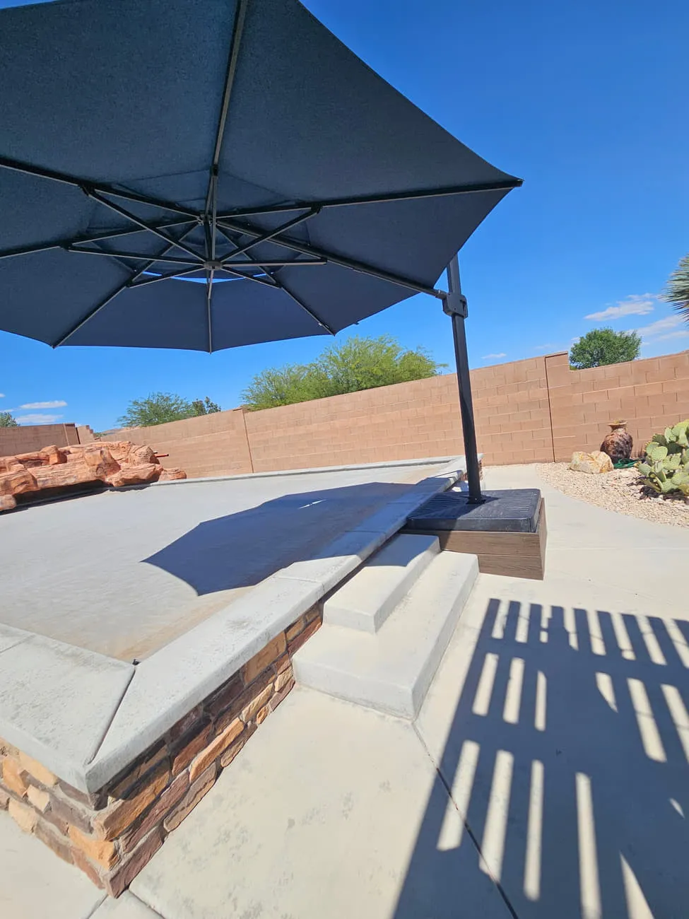 A finished concrete patio with formed steps and a stone-veneer faced raised slab edge, under an open navy cantilever umbrella
