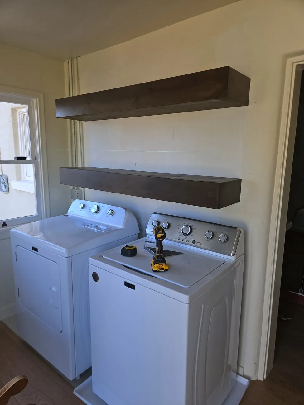 Two chunky dark-stained floating shelves mounted above a white washer and dryer with a laser level line visible on the wall