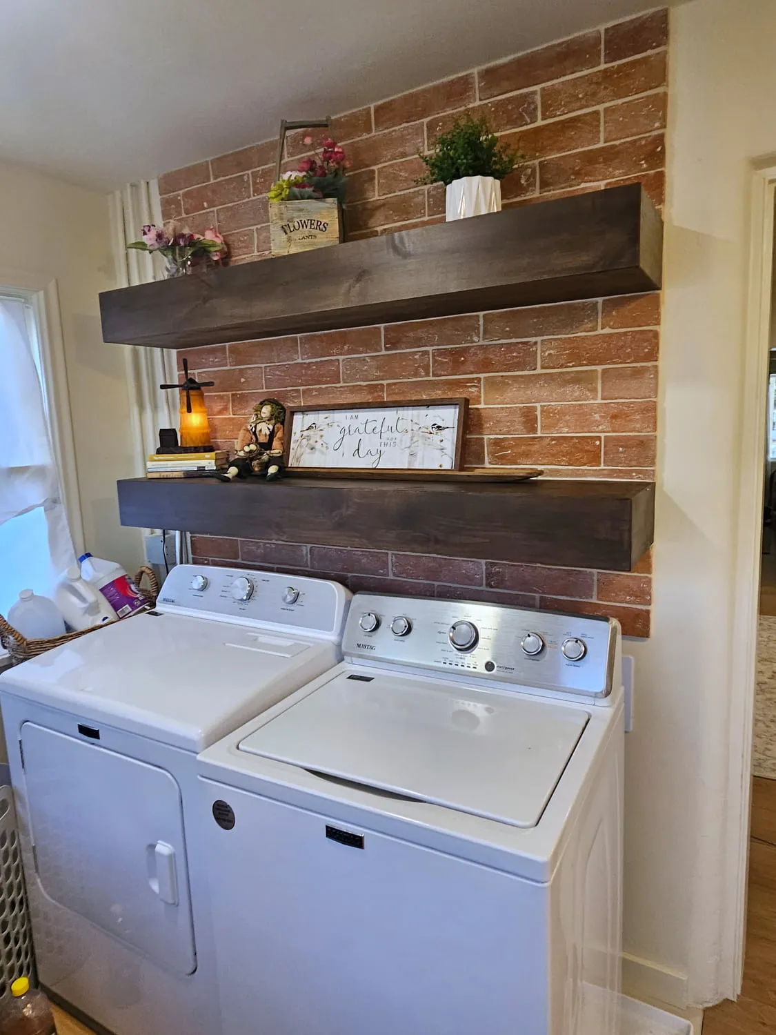 Two stained floating shelves mounted on a hand-laid thin brick accent wall above a white washer and dryer