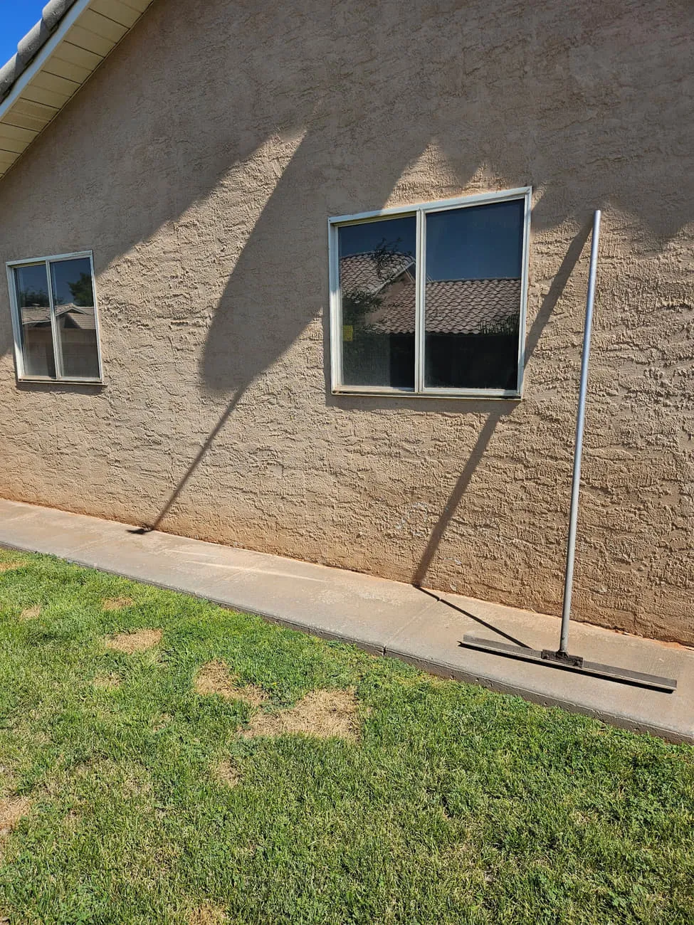 A freshly finished concrete mow strip and sidewalk running along a tan stucco wall with a bull float resting on it