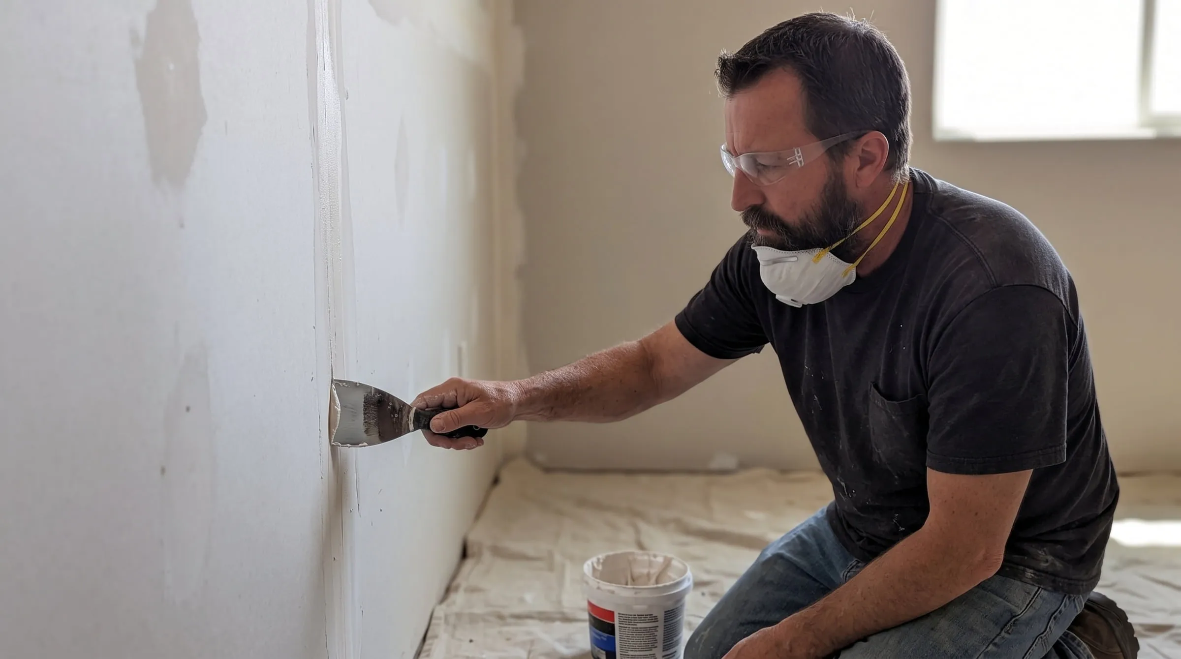 A tradesman in a dust mask feathering joint compound across a wall seam with a wide taping knife