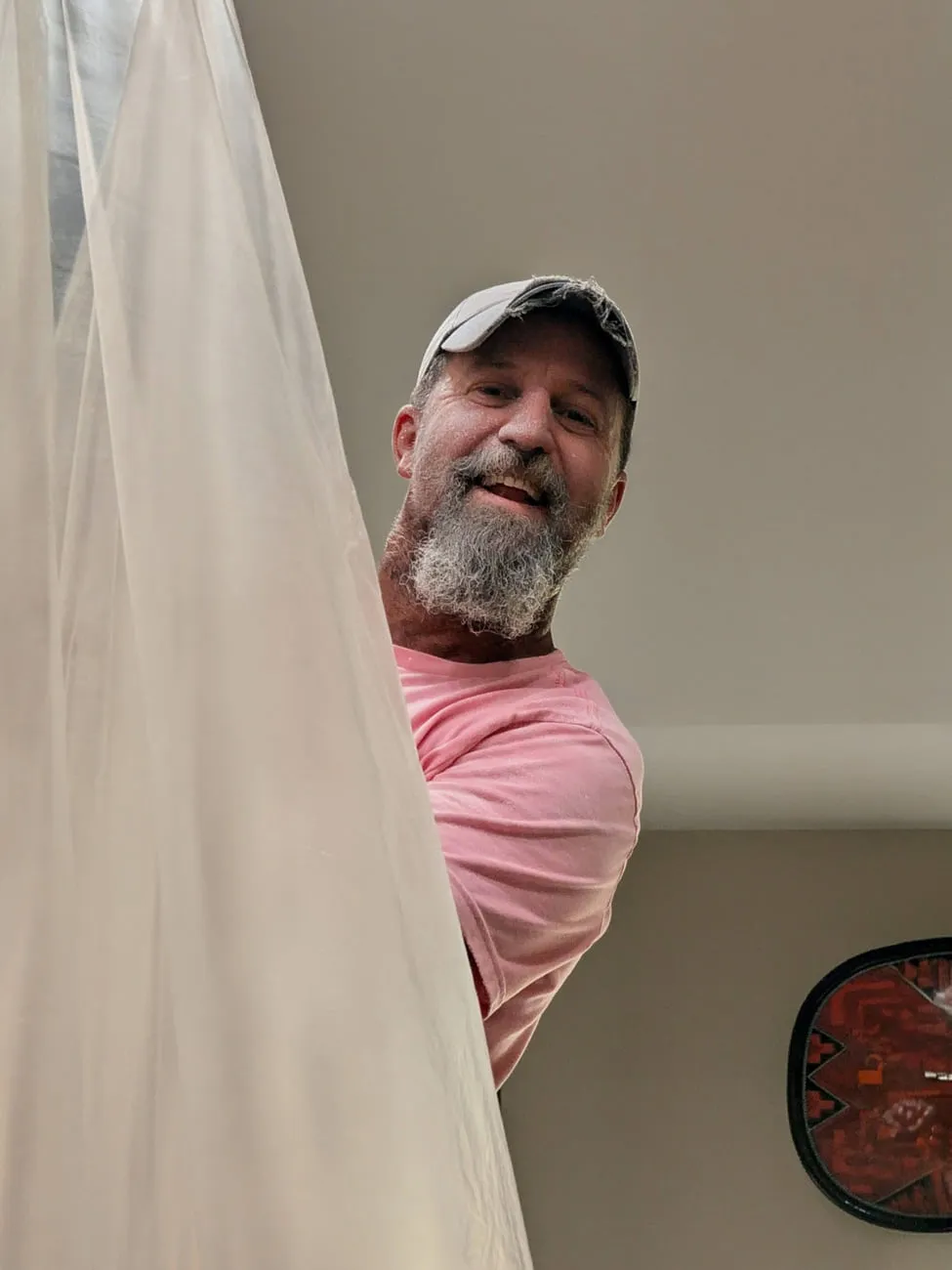 Joe Denter, a grey-bearded man in a white cap and a pink work shirt, grinning around the edge of a hanging dust sheet on a jobsite