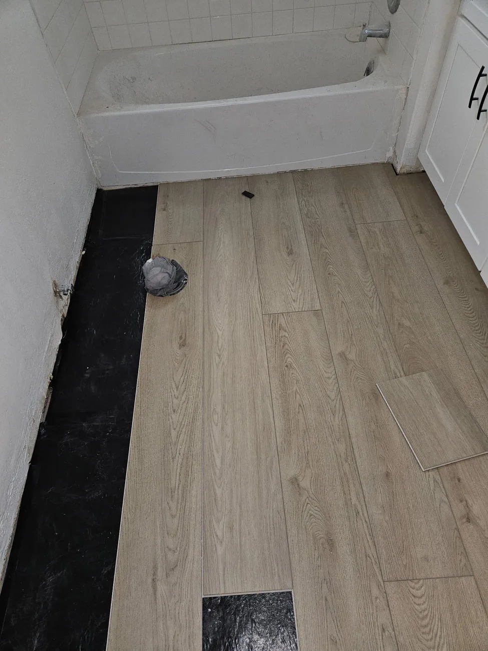 Tan-oak laminate plank part-installed over black underlayment in a bathroom beside an alcove tub